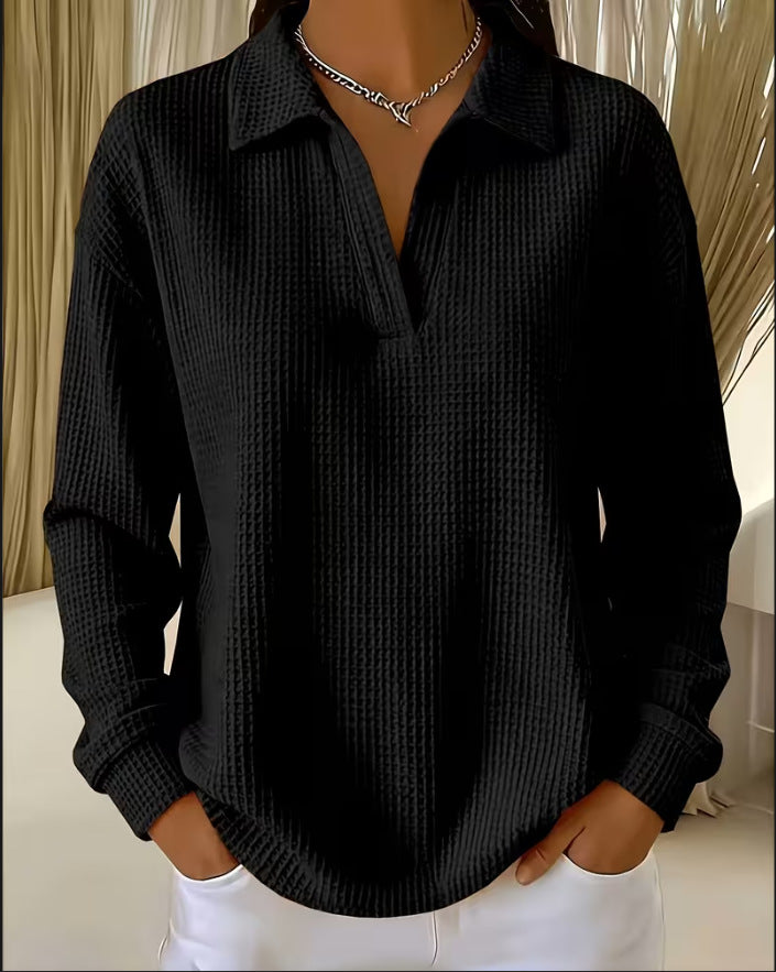 A woman wearing a black long-sleeve waffle-knit collared shirt with a split V-neck, paired with white pants and a silver chain necklace.