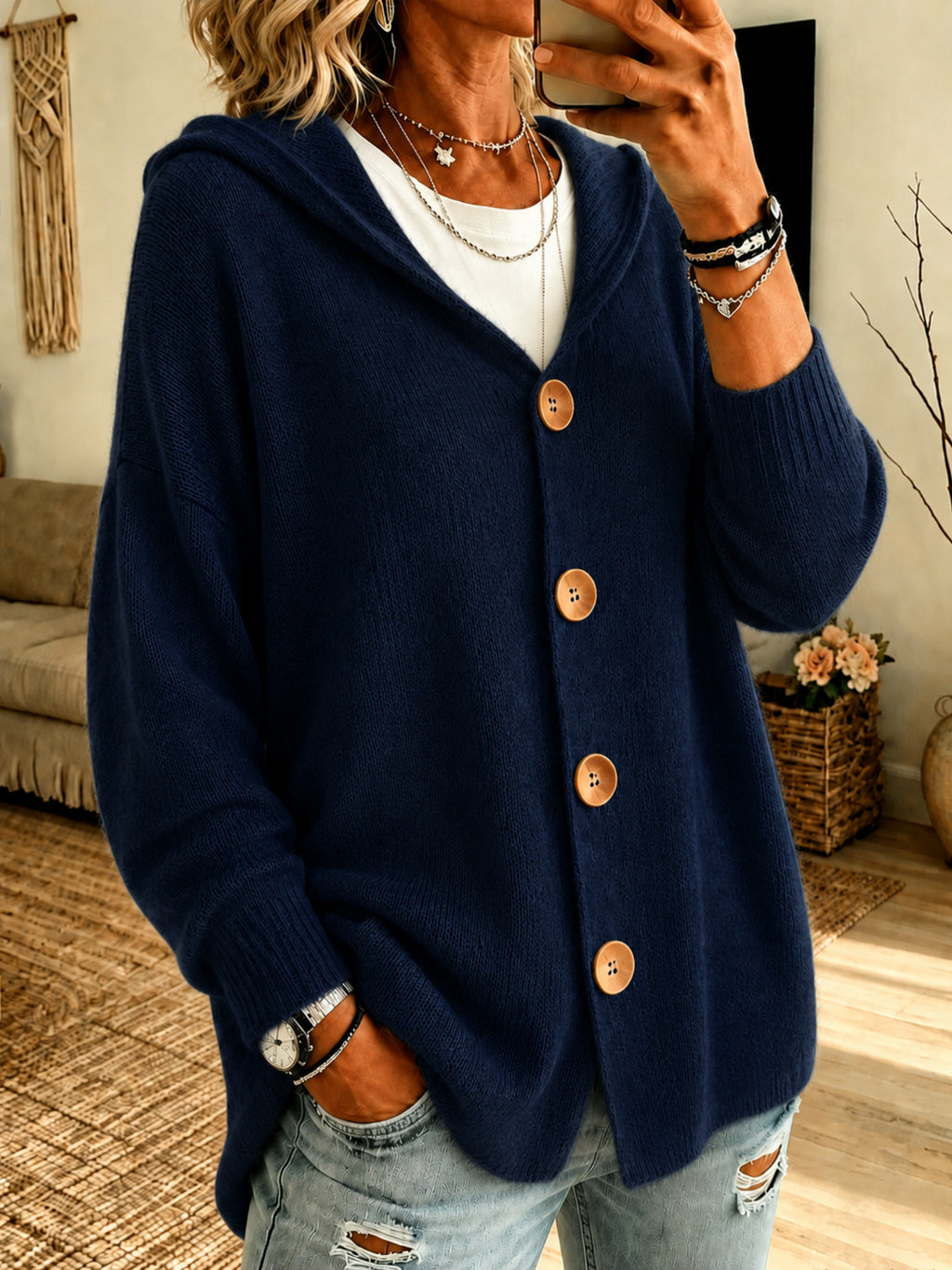 A woman wearing a navy blue knit cardigan with large brown buttons, layered over a white top and paired with distressed light blue jeans in a cozy indoor setting.