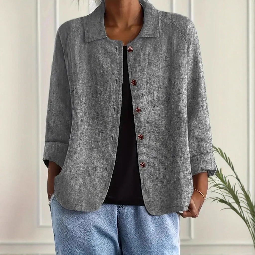 Person wearing a gray jacket over a black top and blue jeans against a white wall.
