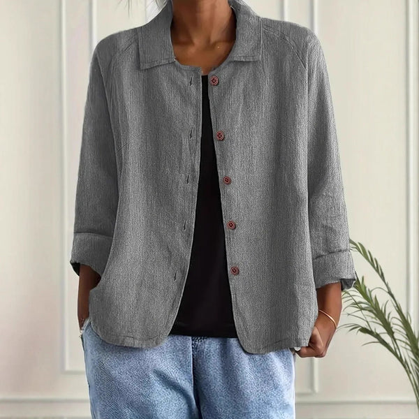 Person wearing a gray jacket over a black top and blue jeans against a white wall.