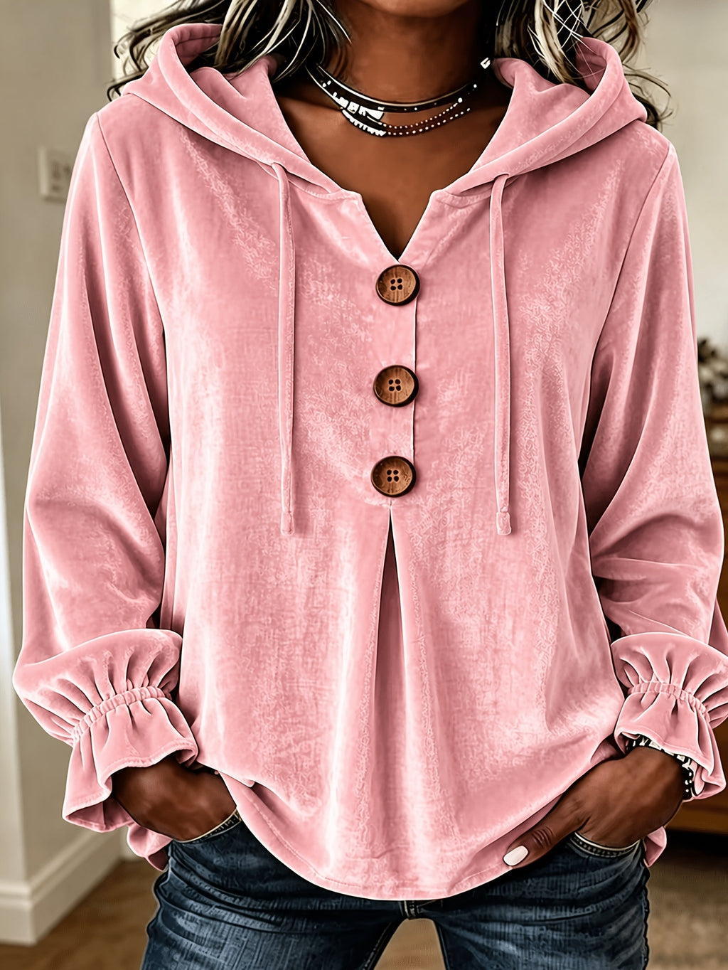 A woman wearing a pink velour hooded long-sleeve blouse with three brown buttons down the front, elasticized ruffled cuffs, and dark blue jeans.