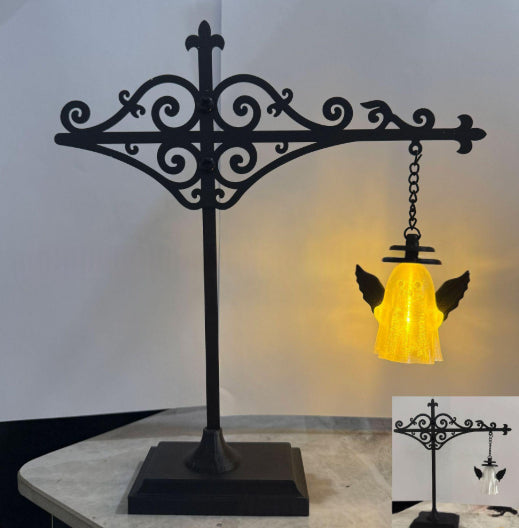 Black wrought iron angel wing hanging lamp stand with a glowing yellow bell-shaped glass shade, shown on a tabletop against a plain wall, with a small inset showing the unlit product.