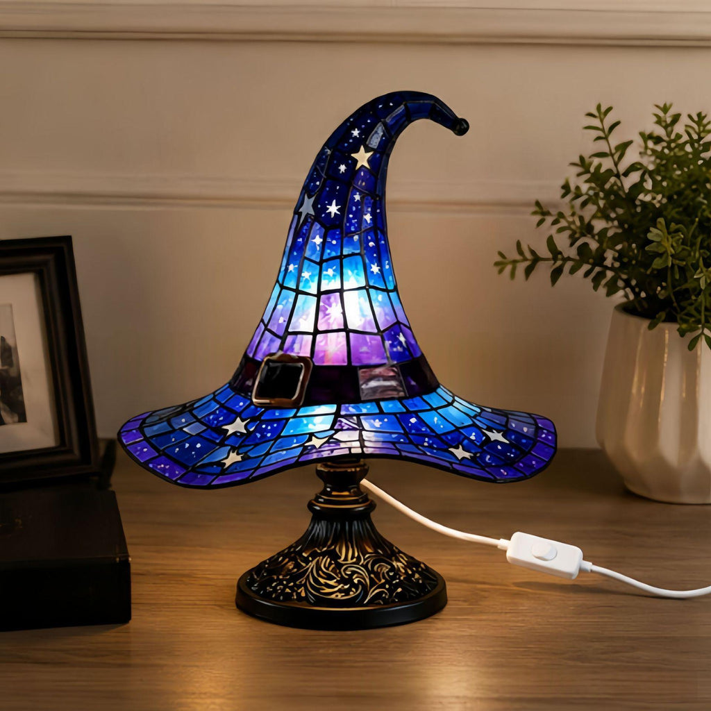 A stained-glass-style witch hat table lamp with a blue, purple, and black galaxy pattern, illuminated on a wooden tabletop with a power cord visible.