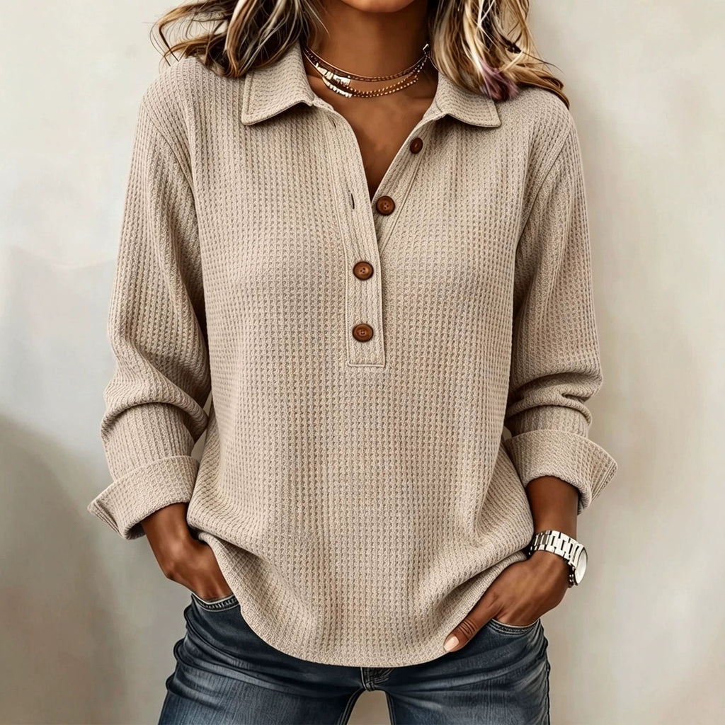 A woman wearing a beige waffle-knit long-sleeve polo shirt with brown buttons, paired with blue jeans, standing against a neutral beige wall.