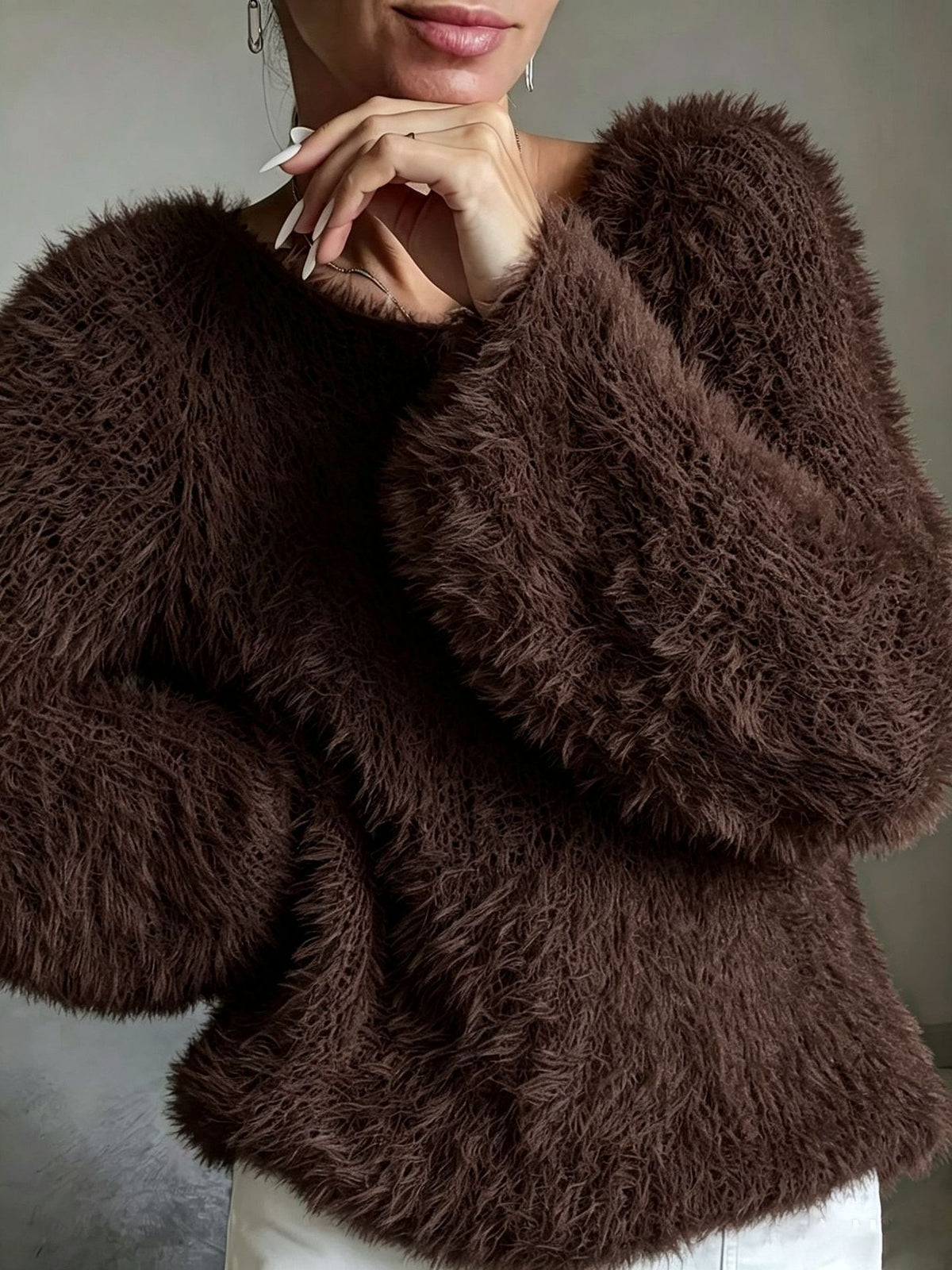 A close-up of a person wearing a dark brown, shaggy faux-fur cropped sweater with voluminous long sleeves, paired with white pants against a neutral gray background.