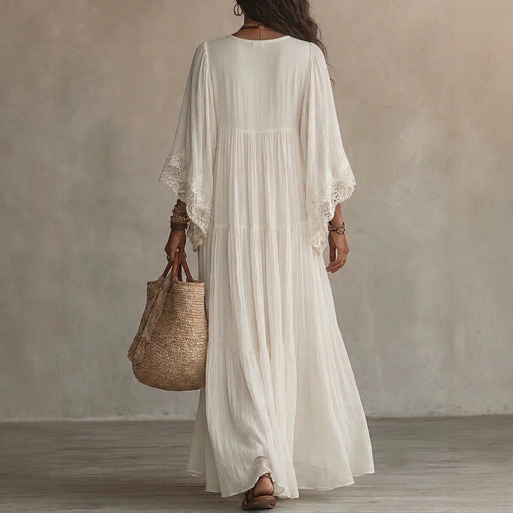A woman viewed from behind wearing a flowing white maxi dress with wide lace-trimmed sleeves, holding a woven straw tote bag against a neutral textured studio backdrop.