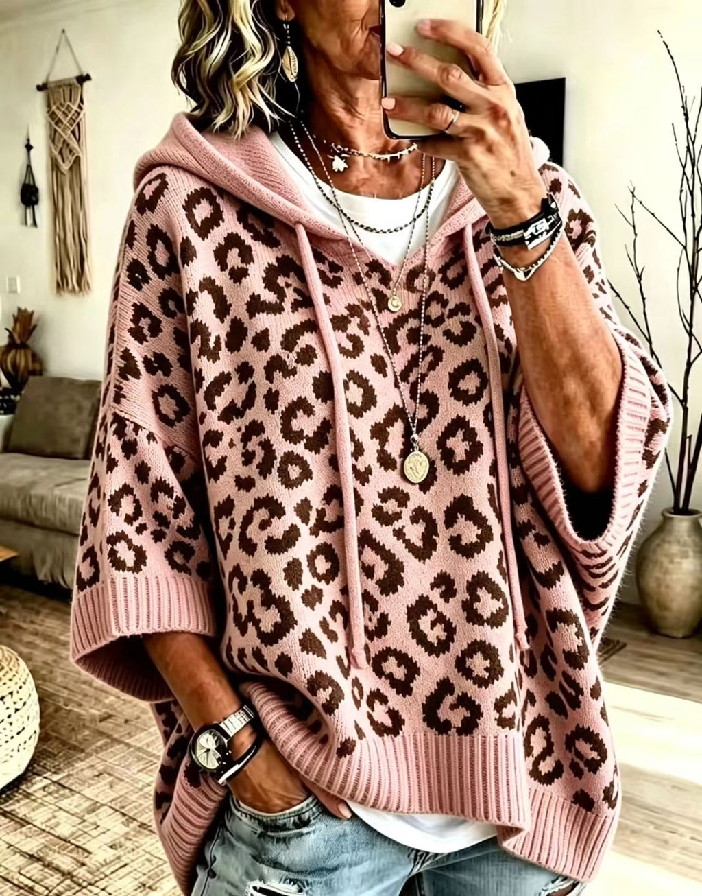 A woman takes a mirror selfie in a cozy living room while wearing a pink leopard-print knit hoodie poncho sweater with brown spots, layered over a white t-shirt and paired with distressed blue jeans.