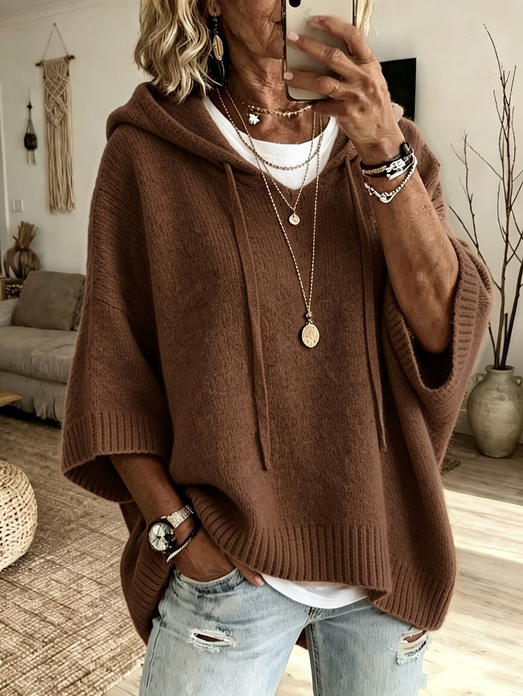 A woman takes a mirror selfie wearing a brown knit poncho hoodie sweater, layered over a white t-shirt and paired with light-wash distressed jeans. She is accessorized with layered gold necklaces, bracelets, and a watch.