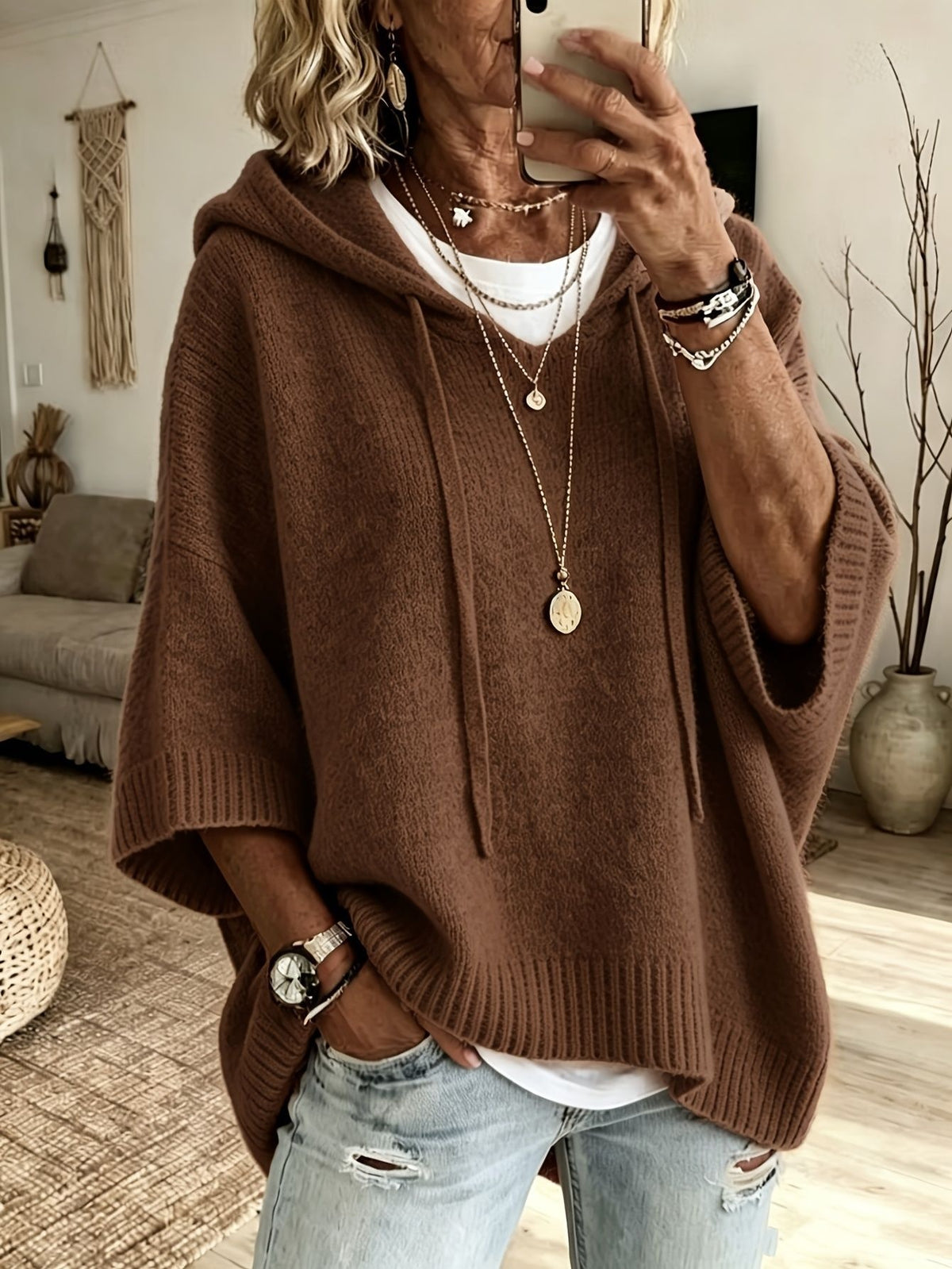 A woman takes a mirror selfie wearing a brown knit poncho hoodie sweater, layered over a white t-shirt and paired with light-wash distressed jeans. She is accessorized with layered gold necklaces, bracelets, and a watch.