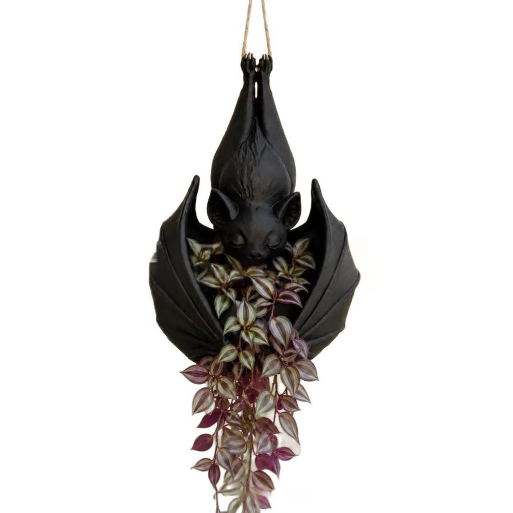 A hanging bat-shaped planter in matte black with a faux trailing plant featuring silver and burgundy leaves, suspended against a plain white background.
