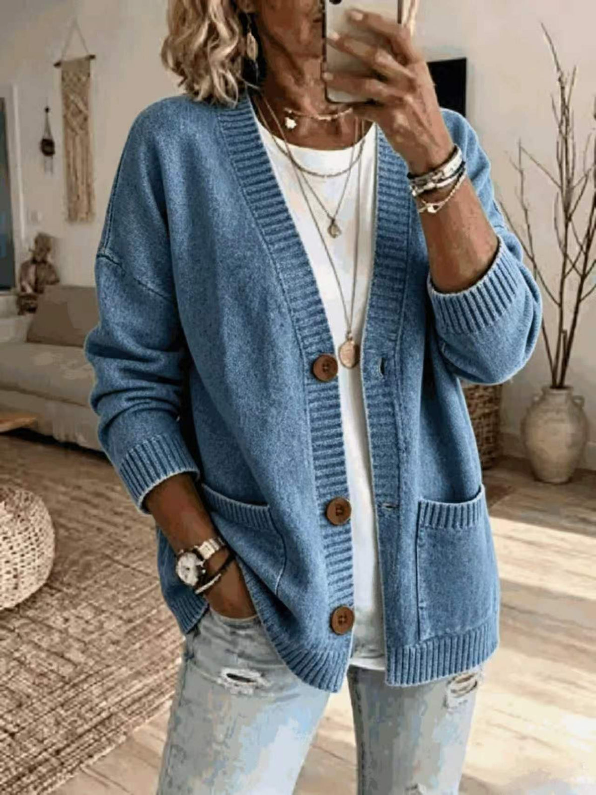 A woman takes a mirror selfie wearing a light blue knit cardigan with brown buttons over a white t-shirt and light-wash distressed jeans, accessorized with layered necklaces and bracelets.
