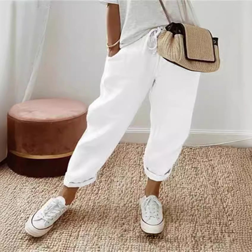 Person wearing white sneakers and white pants with a beige bag, standing on a carpeted floor.