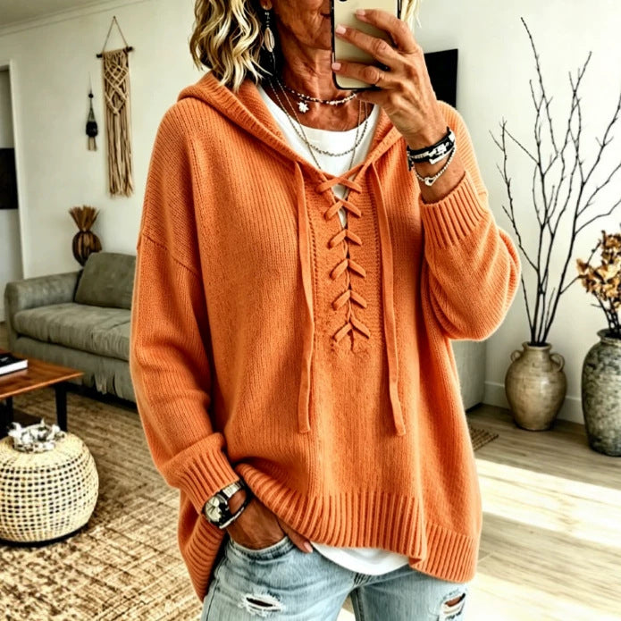 A woman takes a mirror selfie wearing a loose, oversized orange knit hooded sweater with lace-up front detailing, paired with light blue distressed jeans and layered necklaces, set in a cozy living room environment.