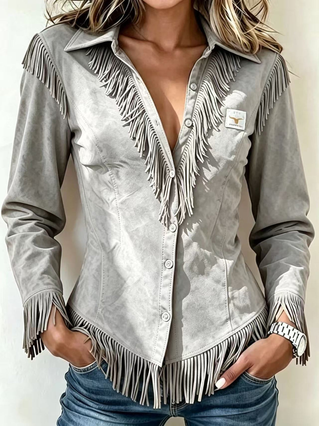 A woman wearing a light gray suede long-sleeve western-style shirt jacket with fringe detailing, paired with blue jeans.