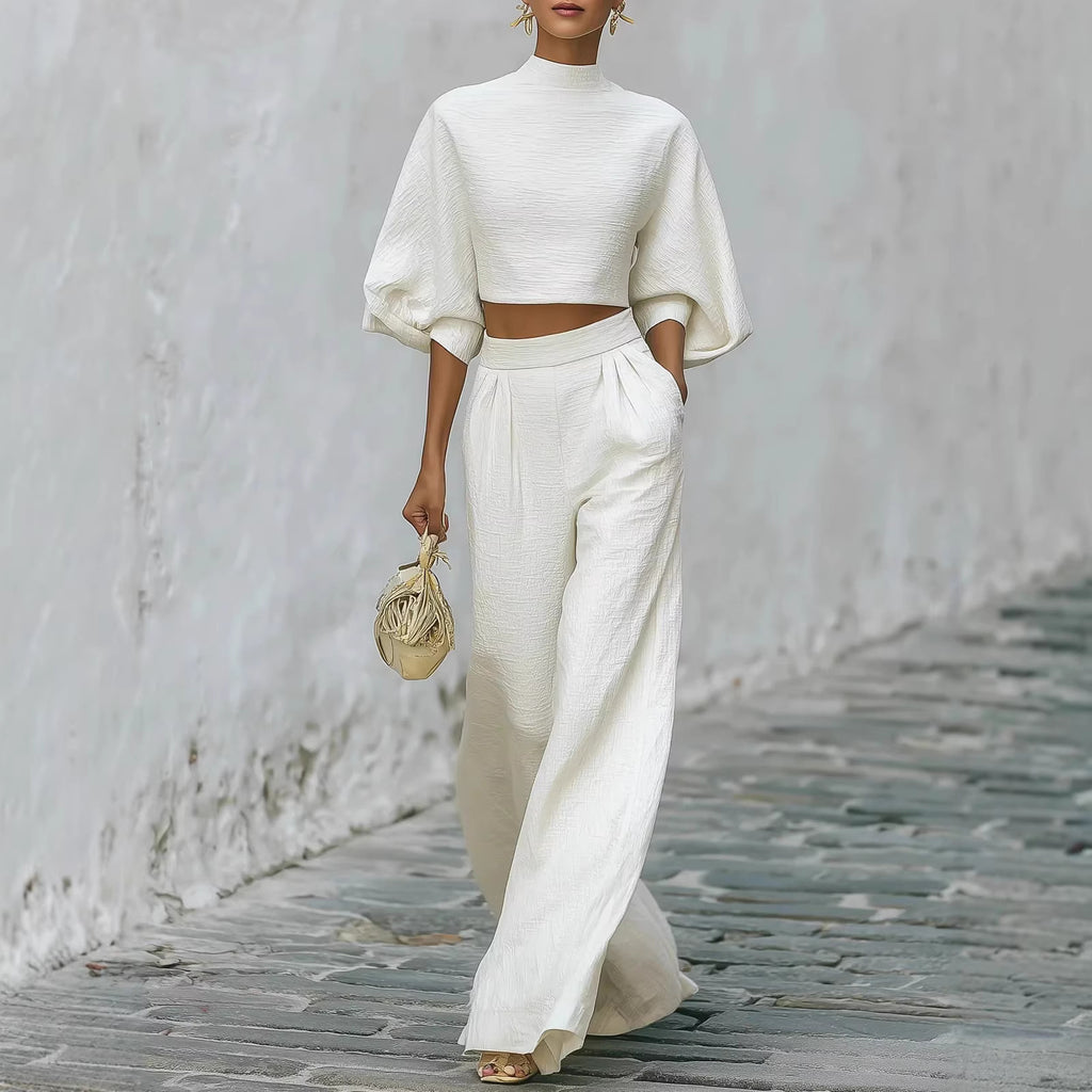 Person wearing a white outfit with wide-legged pants and a crop top, holding a small bag on a stone path.