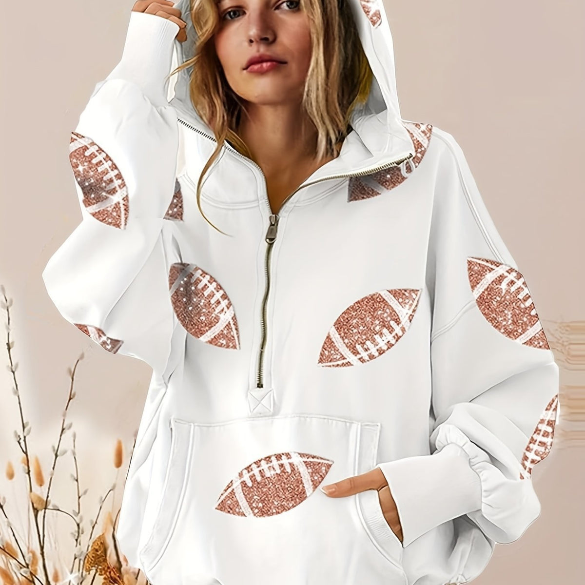 A woman wearing a white half-zip hoodie with rose gold glitter football prints on the sleeves, shoulders, and front pocket, set against a soft beige background.