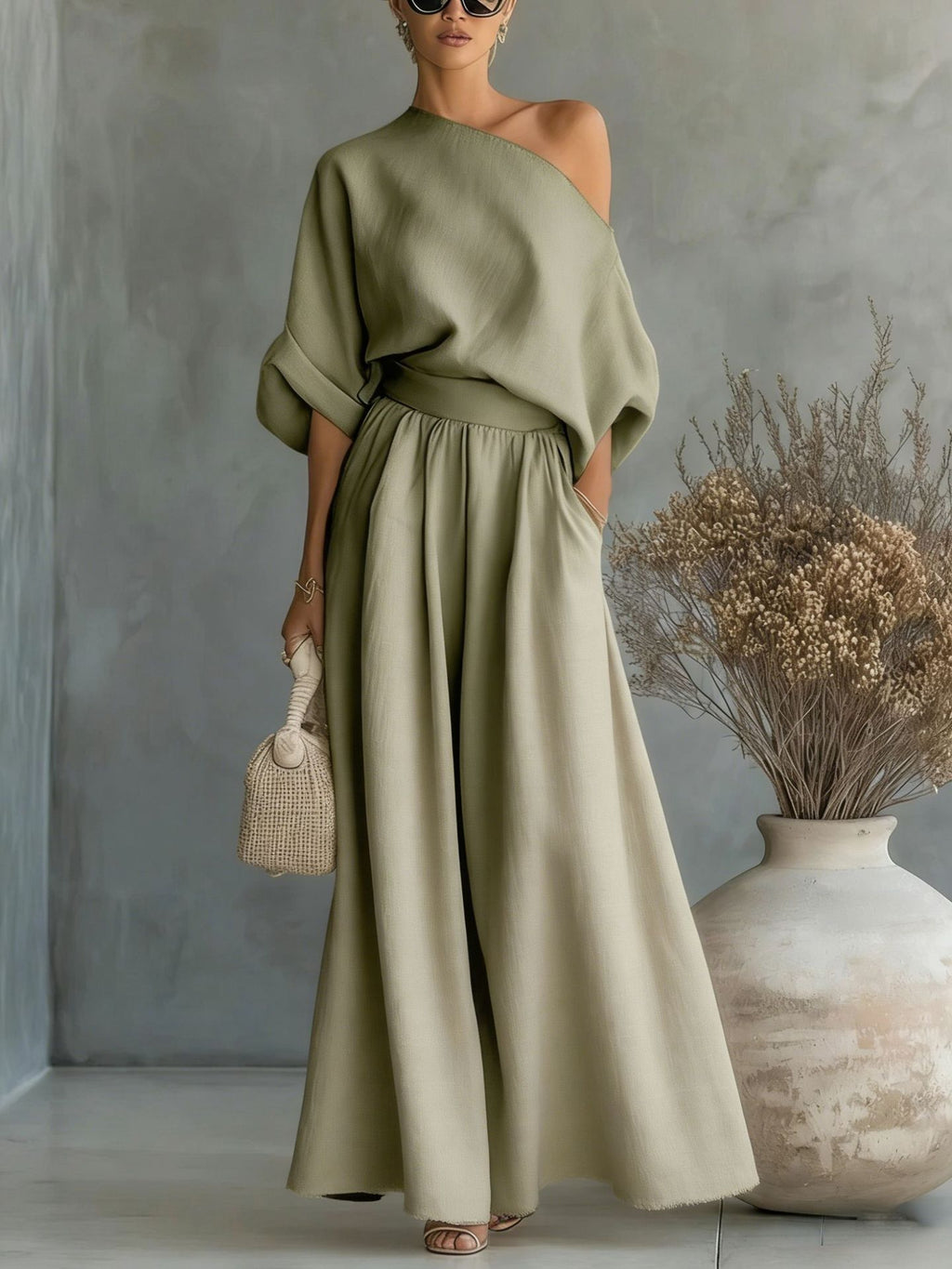 Woman wearing a green off-shoulder top and long skirt standing against a gray wall with a vase and dried plants.