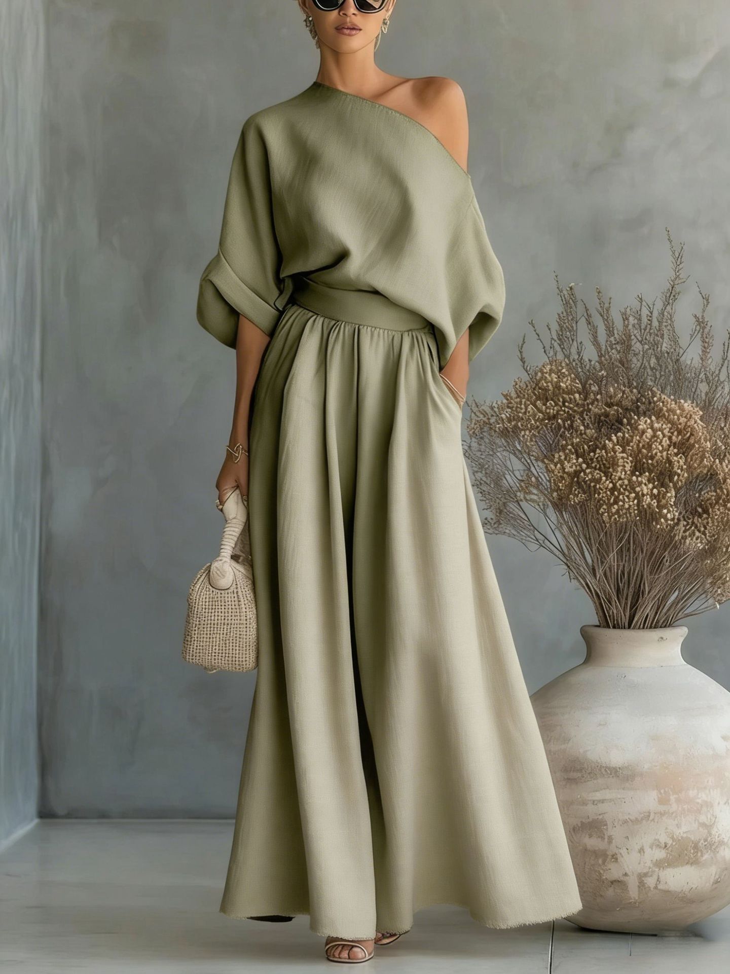 Woman wearing a green off-shoulder top and long skirt standing against a gray wall with a vase and dried plants.