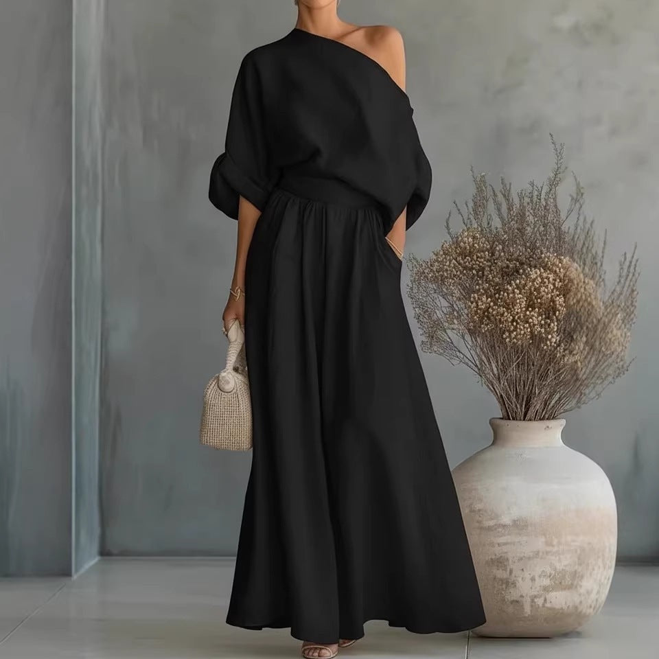 Woman in a black one-shoulder dress holding a small bag, standing next to a textured vase with dried plants.