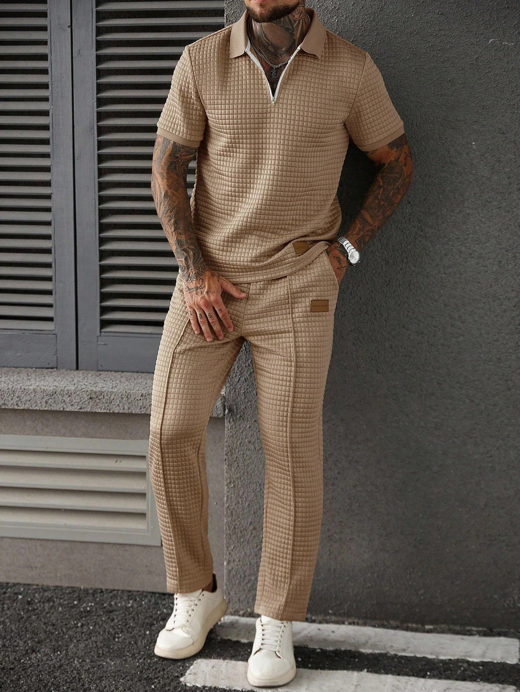 Man wearing a beige checkered outfit with white shoes against a gray wall.