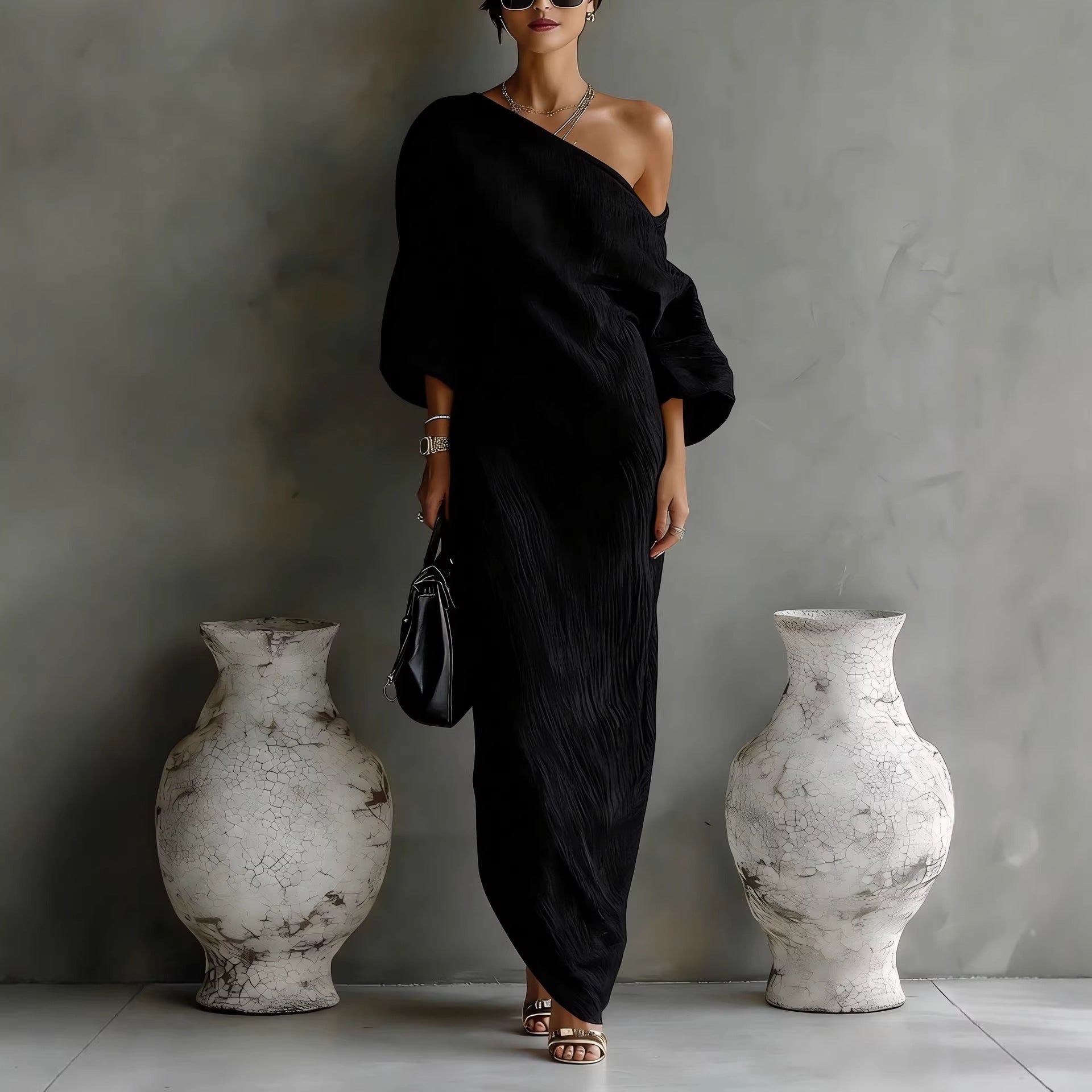 Woman in a black off-shoulder dress standing between two large white vases against a gray wall.