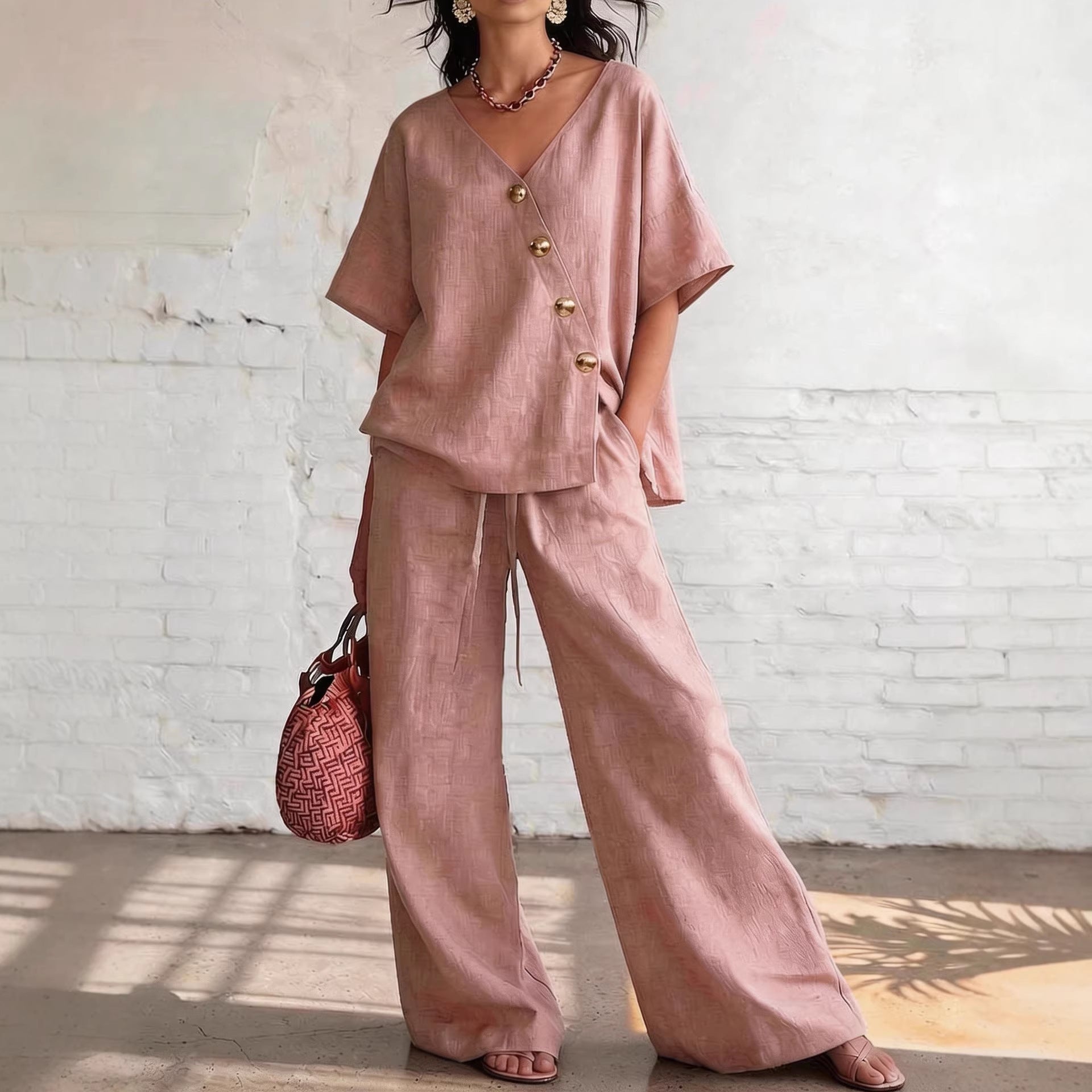 Woman wearing a pink outfit with wide-leg pants and a top with gold buttons, holding a red bag against a white brick wall.