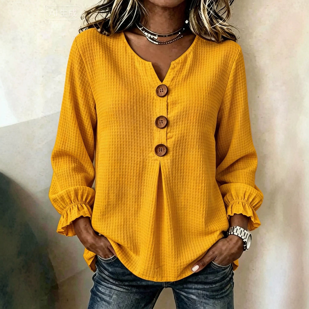 A woman wearing a mustard yellow waffle-knit long-sleeve blouse with a split neckline, three brown buttons, and ruffled cuffs, paired with dark blue jeans.
