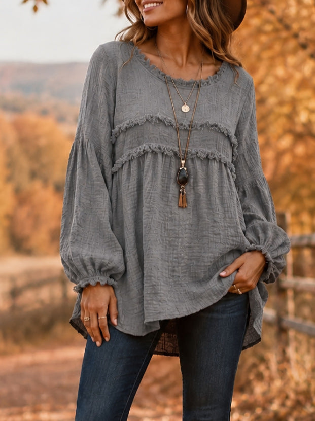 A woman wearing a gray long-sleeve blouse with layered ruffle details and elastic cuffs, paired with dark blue jeans, standing outdoors in a blurred autumn landscape.