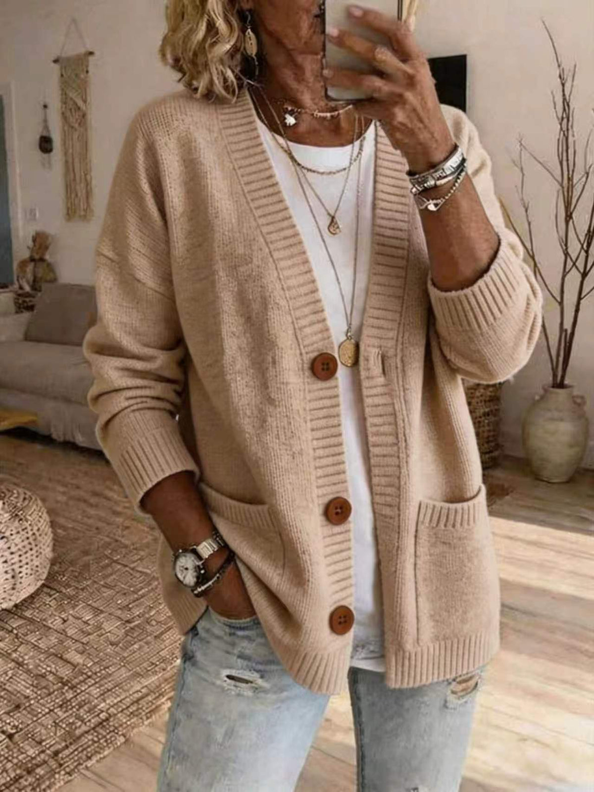 A woman takes a mirror selfie wearing a beige open-front knit cardigan with brown buttons over a white top and light blue jeans, in a cozy indoor room setting.
