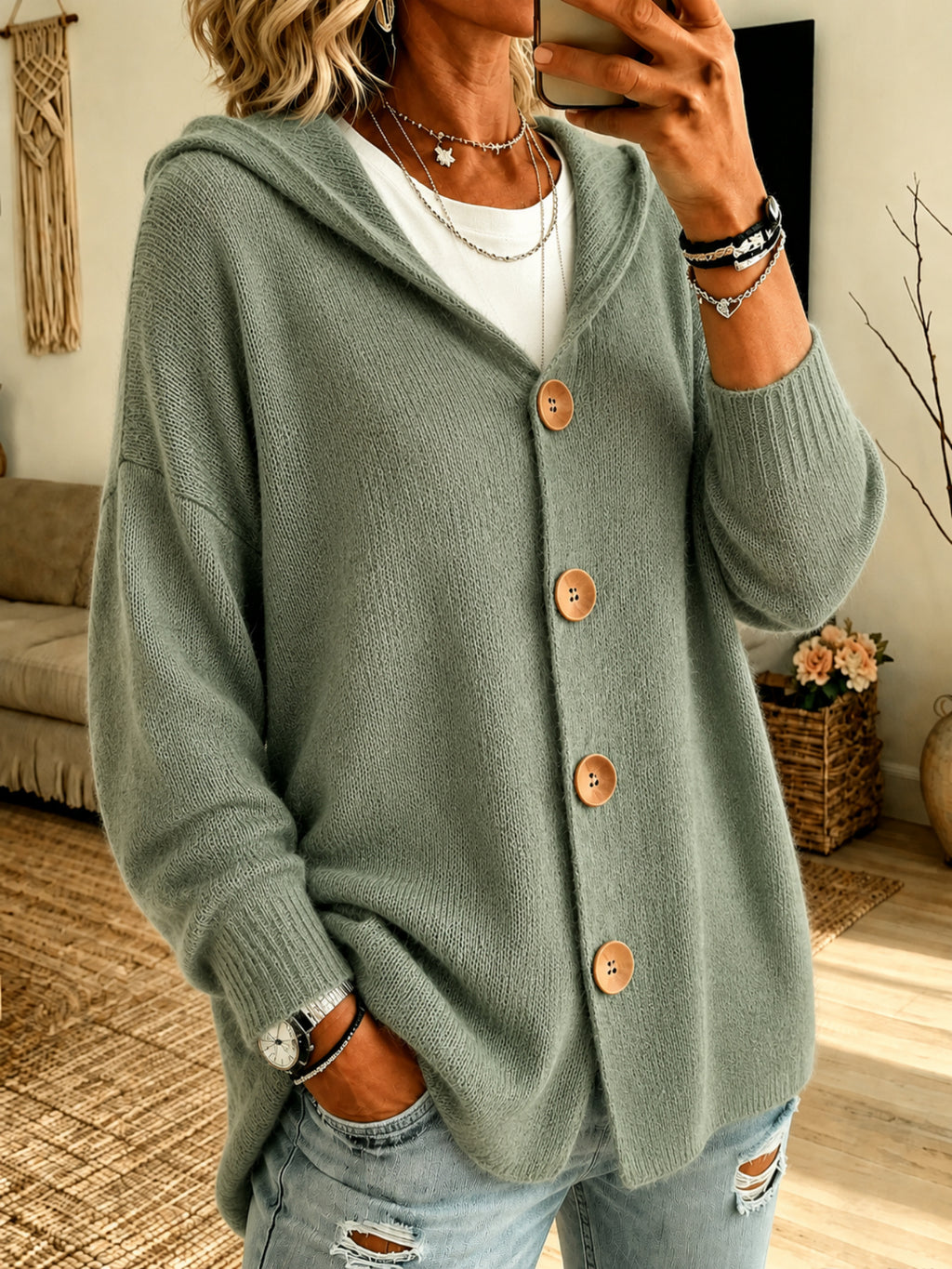 A woman wearing a sage green knit cardigan with a hood and large brown buttons, layered over a white top and paired with distressed light blue jeans in a cozy indoor setting.