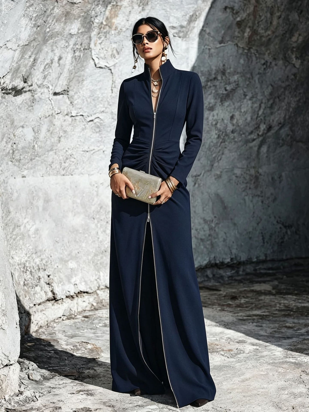 A woman stands outdoors against a textured stone wall wearing a long navy maxi dress with a high collar, front zipper, ruched waist, and wide legs. She is holding a metallic clutch and wearing sunglasses and jewelry.