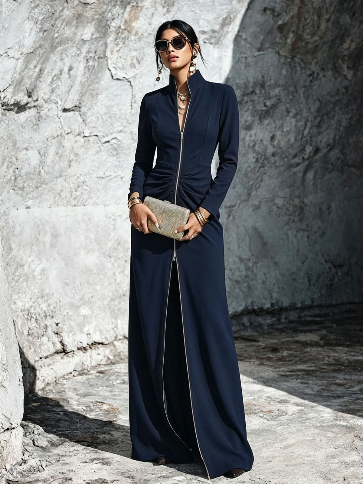 A woman stands outdoors against a textured stone wall wearing a long navy maxi dress with a high collar, front zipper, ruched waist, and wide legs. She is holding a metallic clutch and wearing sunglasses and jewelry.