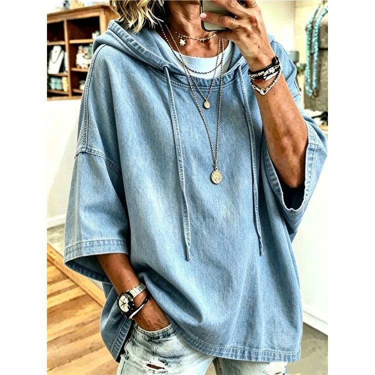 A woman wearing an oversized light blue denim hooded sweatshirt with drawstrings, layered with multiple gold necklaces and bracelets, taking a mirror selfie in an indoor room with wooden floors and shelves.