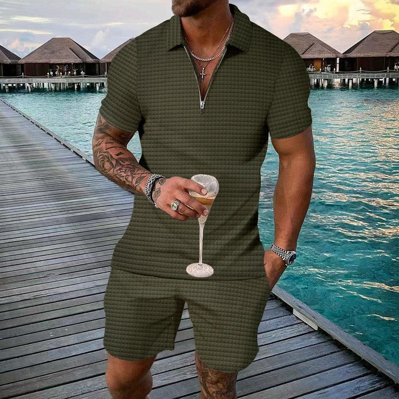Man in green checkered outfit holding a drink on a dock with water and buildings in the background