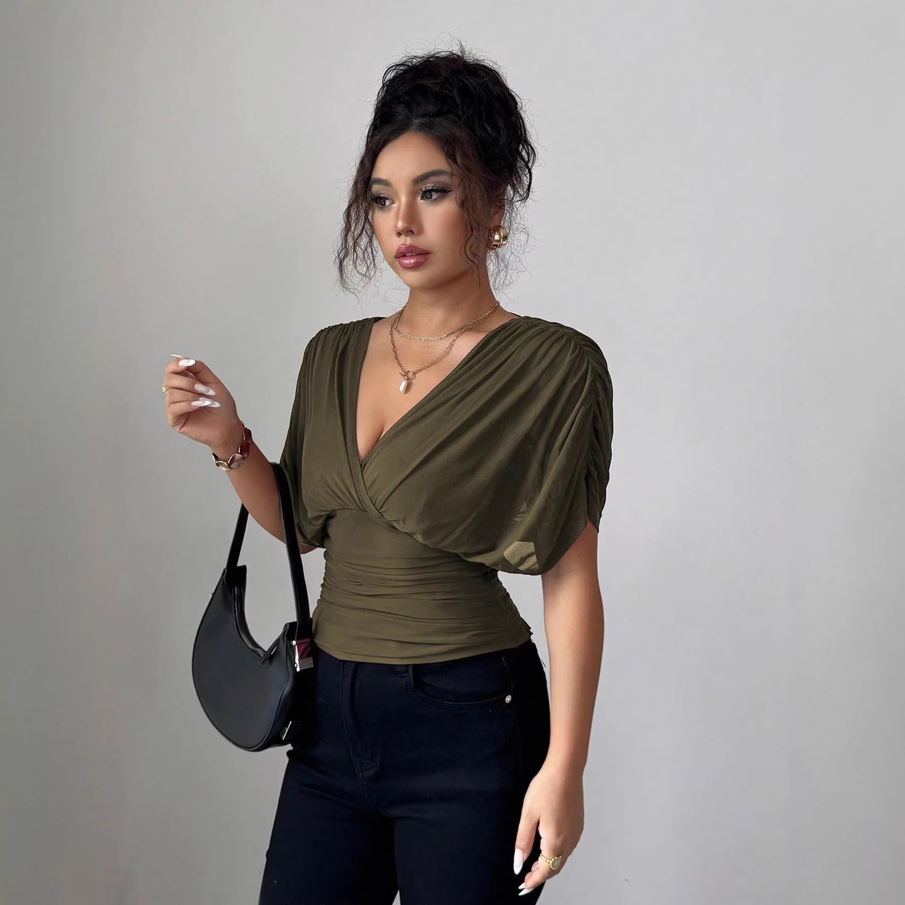 Woman in olive green top and black pants holding a black handbag against a gray background