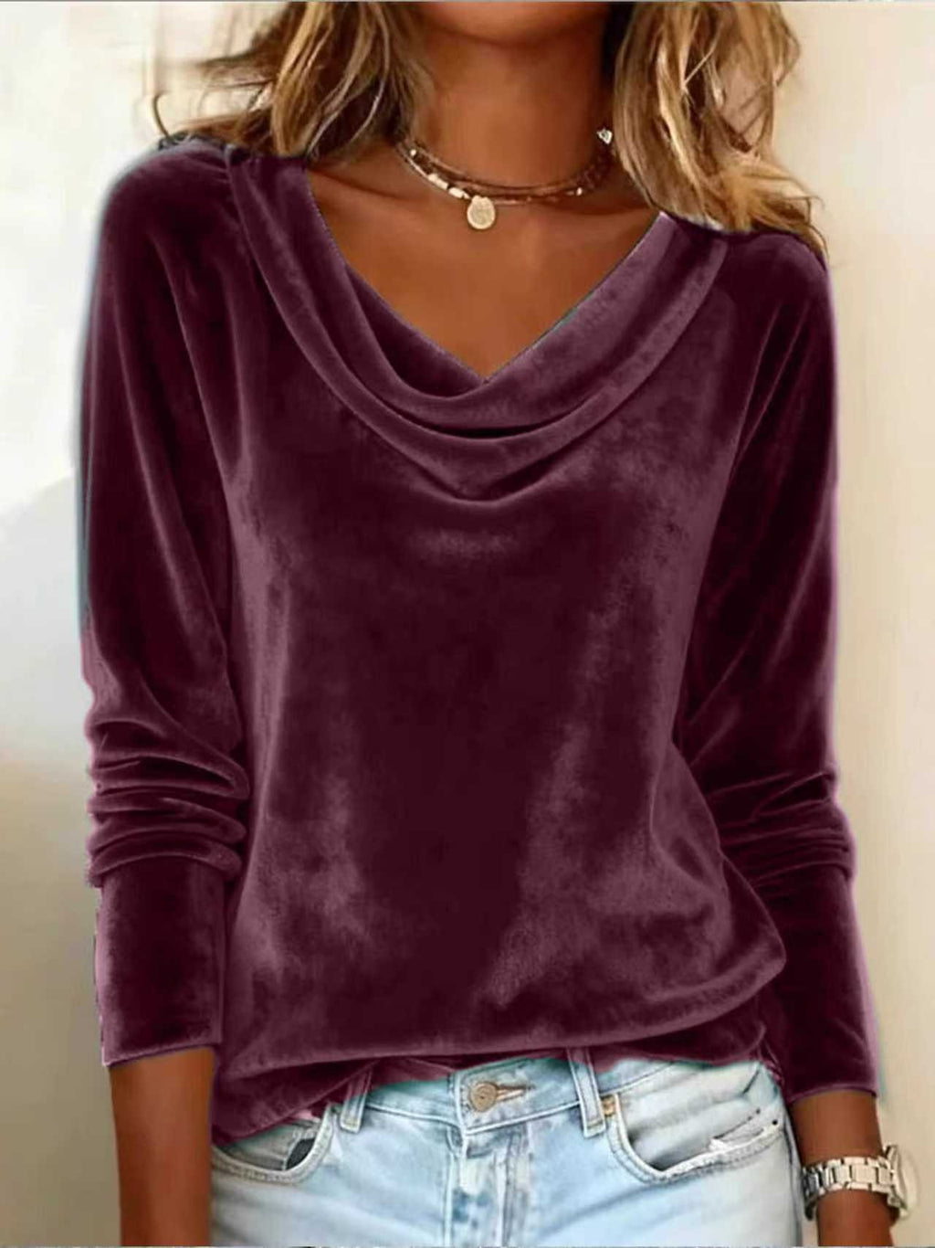 A woman wearing a long-sleeve cowl neck velvet top in deep plum purple, paired with light blue jeans and layered gold necklaces.