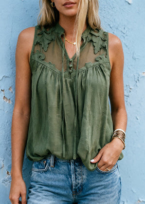A woman wearing an olive green sleeveless blouse with sheer floral lace panels and a gathered front, paired with blue jeans, standing against a textured light blue wall.