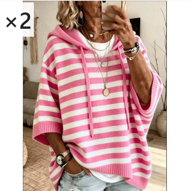 A woman takes a mirror selfie wearing a pink and white horizontally striped knit hoodie with wide three-quarter sleeves and a drawstring hood. She is also wearing layered gold necklaces, bracelets, and a watch, paired with light-wash denim jeans.