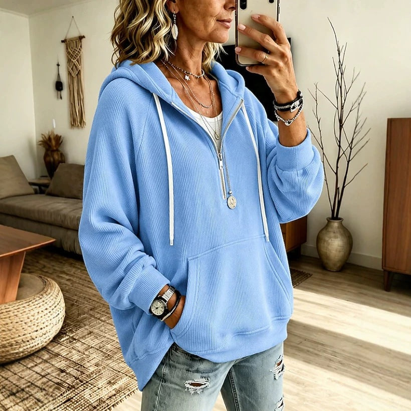 A woman takes a mirror selfie wearing a light blue ribbed zip-up hoodie with white drawstrings and a front kangaroo pocket, paired with distressed light-wash jeans in a modern living room setting.