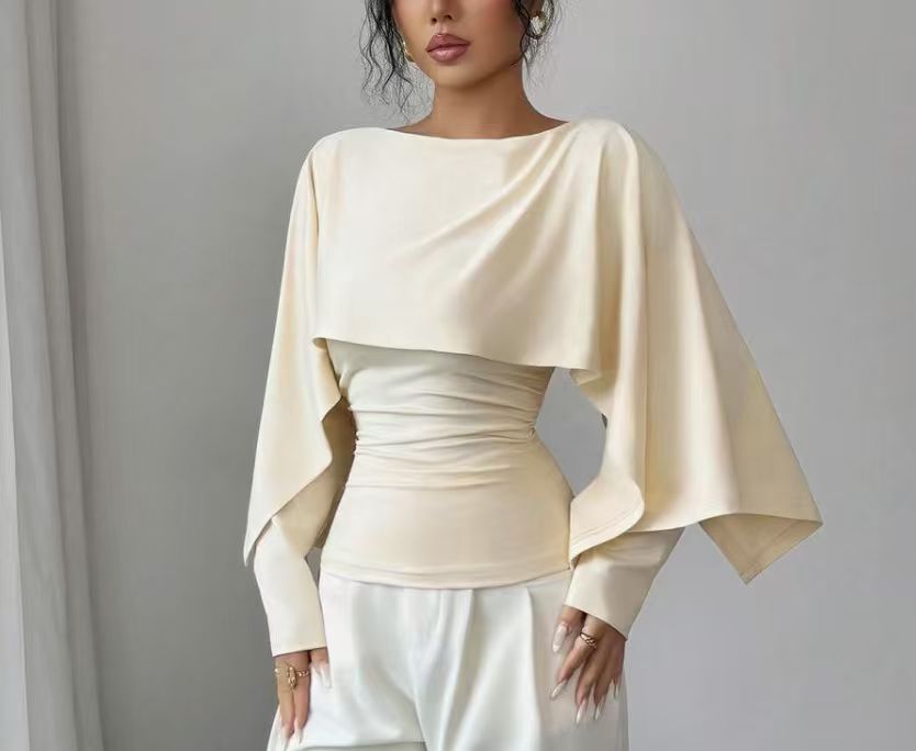 A woman wearing a cream-colored draped long-sleeve top with a ruched waist and white satin pants, standing against a neutral gray background.