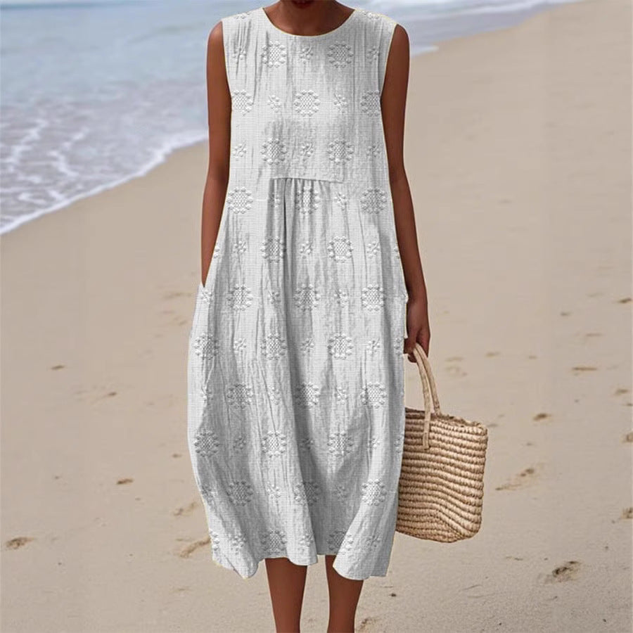 Woman in a white sleeveless dress holding a straw bag on a beach.