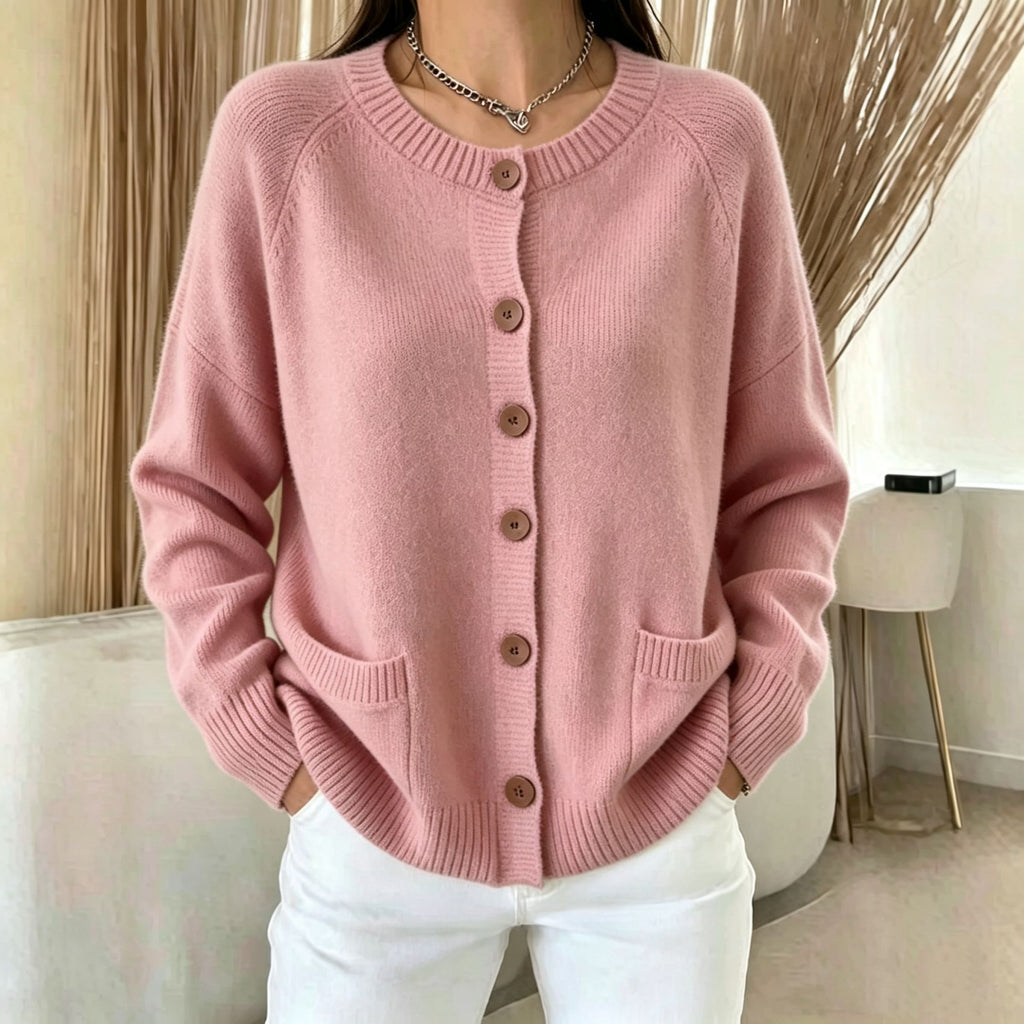 A woman wearing a dusty pink knit cardigan sweater with brown buttons and white pants, standing in a modern indoor setting with neutral-toned curtains and a chair.