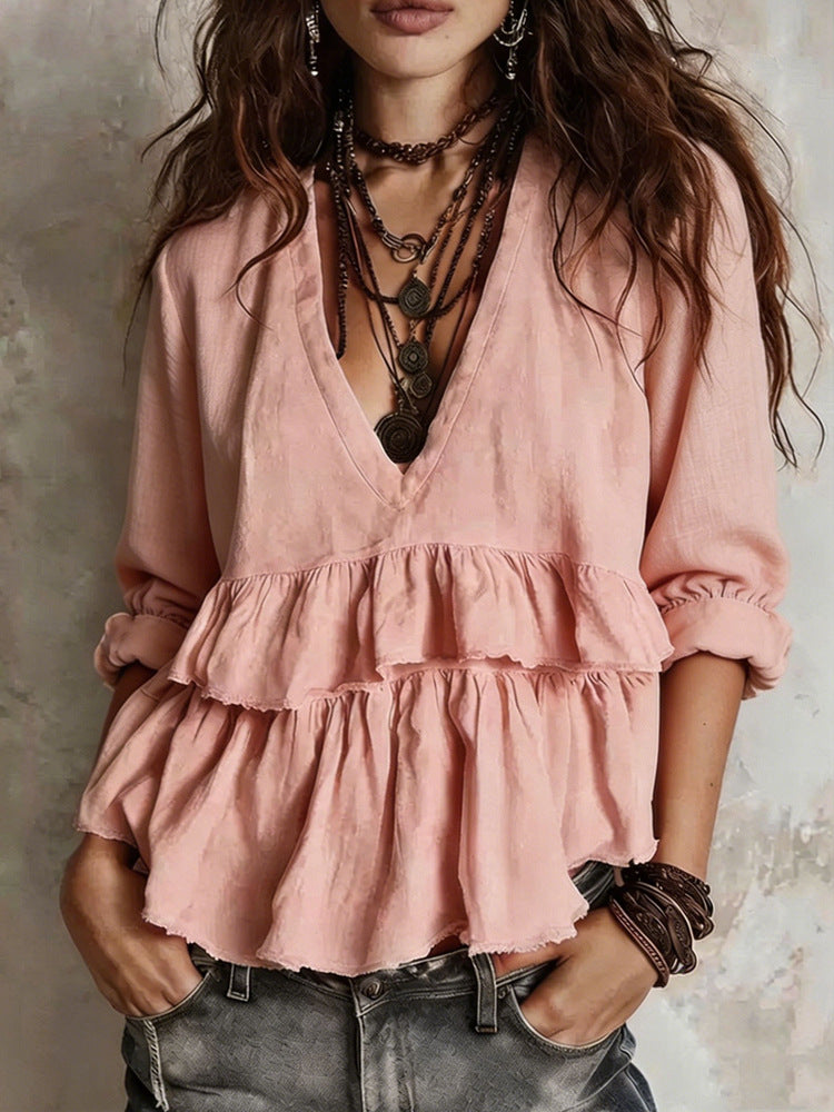A woman wearing a dusty pink bohemian-style blouse with a deep V-neck, layered ruffle details at the waist, and rolled sleeves, paired with dark gray jeans and layered necklaces.