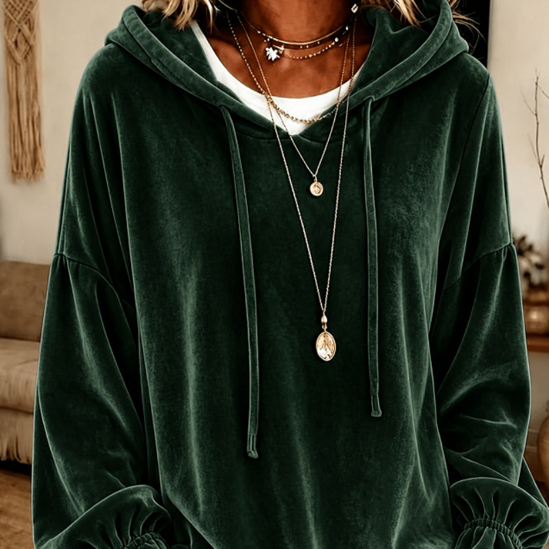 A person wearing an oversized dark green velvet hoodie with drawstrings, layered over a white undershirt and accessorized with multiple gold necklaces, in an indoor room setting.