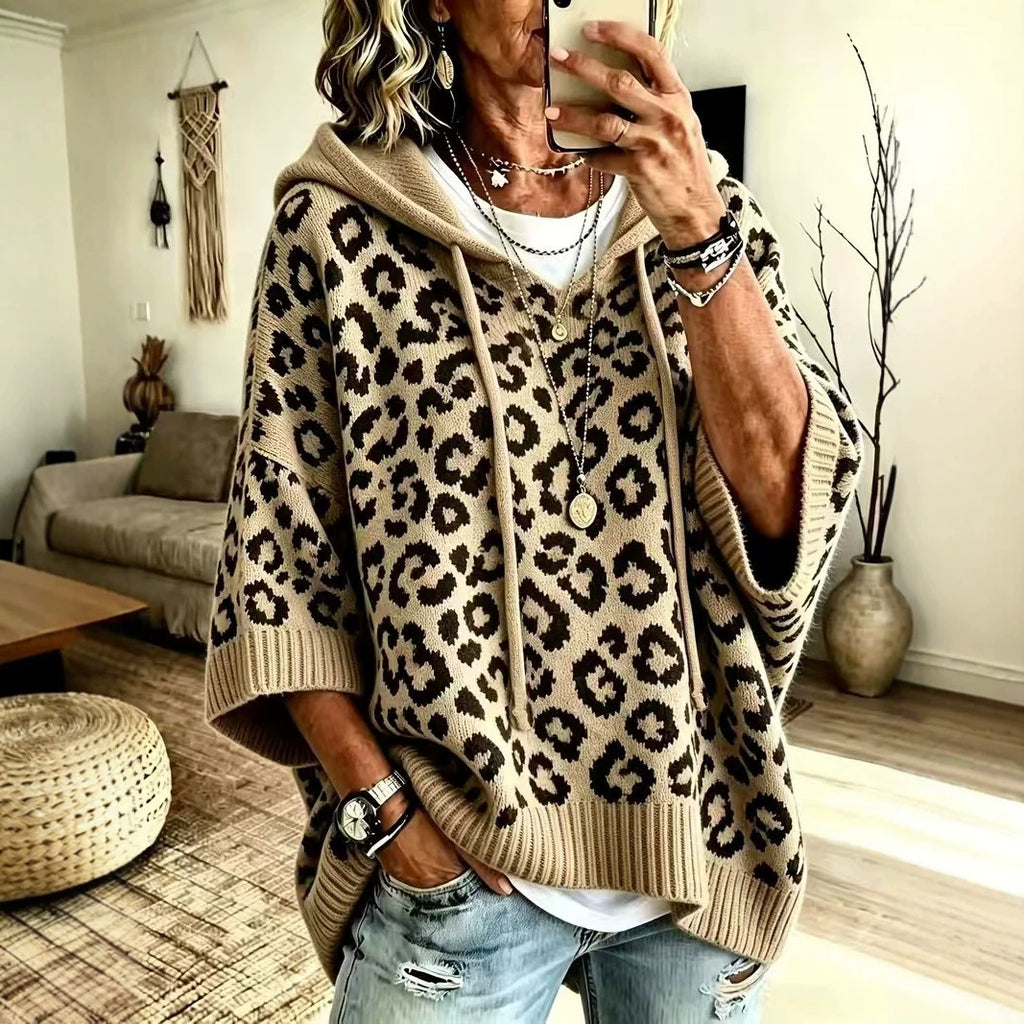 A woman takes a mirror selfie wearing a beige and black leopard-print knit poncho hoodie with a ribbed hem and cuffs, paired with distressed blue jeans in a cozy living room setting.