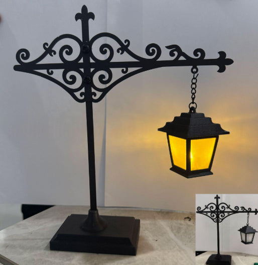 Black wrought iron decorative lamp stand with an ornate scrollwork top and a hanging lantern that glows yellow when lit, shown on a tabletop against a plain light background.