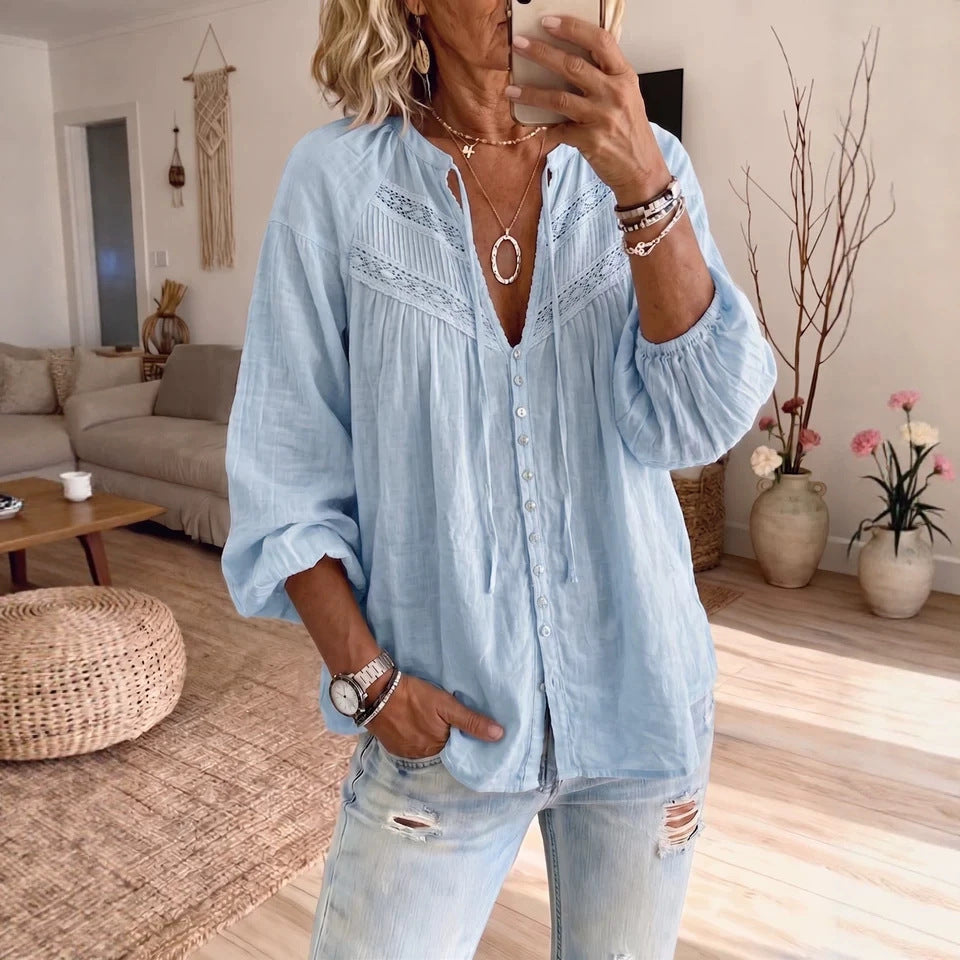 A woman takes a mirror selfie in a cozy living room, wearing a light blue long-sleeve button-up shirt with lace-up front detailing and distressed light-wash jeans.