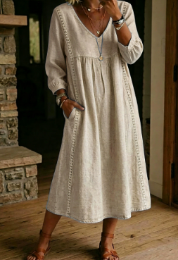 A woman wearing a light beige linen midi dress with long sleeves, a V-neckline, and side pockets, standing in a rustic indoor setting with a stone fireplace and wooden floor.