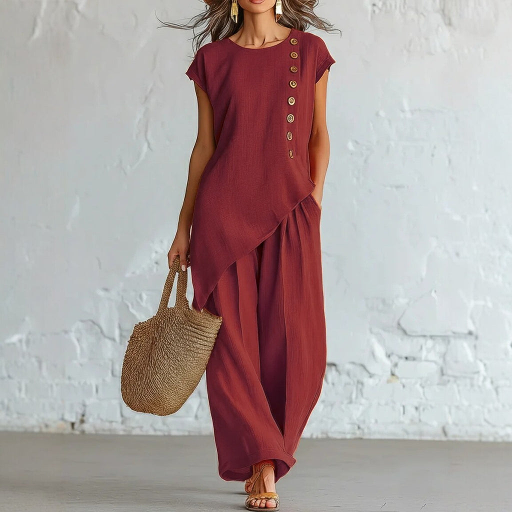 Woman wearing a burgundy outfit with wide-leg pants and a straw bag against a white brick wall.