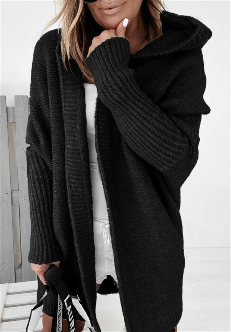A woman wearing a black oversized ribbed knit cardigan with a hood, layered over a white top and white pants, holding a black and white handbag.